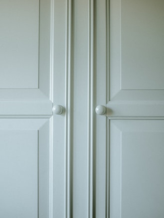 A close-up of a light-colored cabinet door featuring raised panels and two round white knobs. The cabinet's surface has a smooth finish with slight shadows highlighting the contours of the panels.