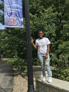 A young woman stands by a black lamppost on a stone path. She is wearing a white t-shirt with the words 'Queens are born in July' and ripped jeans. The background is filled with lush green trees, and there is a banner on the lamppost with the text 'That's the beauty of Hanover' featuring a large 'H' and a panther graphic.