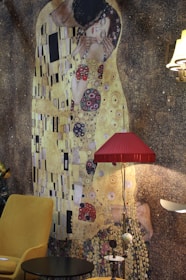 A large mural of a famous painting depicting two figures in an intimate embrace featuring a gold and patterned background, with a modern room setting including a mustard yellow chair, a small black table, a red lampshade, and other furniture pieces creating an eclectic mix of classic and contemporary styles.