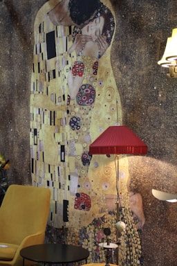 A large mural of a famous painting depicting two figures in an intimate embrace featuring a gold and patterned background, with a modern room setting including a mustard yellow chair, a small black table, a red lampshade, and other furniture pieces creating an eclectic mix of classic and contemporary styles.