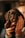 A small black puppy is being held gently by a person's hands. The puppy has soft, fluffy fur and large, soulful eyes. The background is softly blurred, emphasizing the adorable puppy's face.