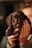 A small black puppy is being held gently by a person's hands. The puppy has soft, fluffy fur and large, soulful eyes. The background is softly blurred, emphasizing the adorable puppy's face.