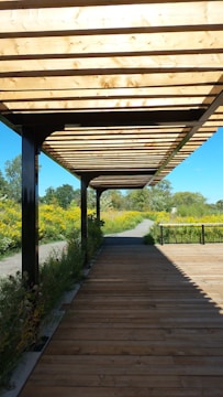 A wooden pergola structure provides shade over a wooden pathway, with lush greenery and yellow flowers flanking the path on either side. The background includes a clear blue sky and distant trees, creating a serene outdoor setting.