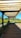 A wooden pergola structure provides shade over a wooden pathway, with lush greenery and yellow flowers flanking the path on either side. The background includes a clear blue sky and distant trees, creating a serene outdoor setting.