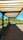A wooden pergola structure provides shade over a wooden pathway, with lush greenery and yellow flowers flanking the path on either side. The background includes a clear blue sky and distant trees, creating a serene outdoor setting.