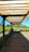 A wooden pergola structure provides shade over a wooden pathway, with lush greenery and yellow flowers flanking the path on either side. The background includes a clear blue sky and distant trees, creating a serene outdoor setting.