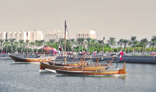 Traditional wooden boats float peacefully on a calm waterfront with palm trees lining the shoreline. Modern buildings with Arabic architectural elements are visible in the background.