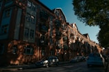 A row of classic brick apartment buildings under a clear blue sky, with shadows from nearby trees casting patterns on the facade. Several cars are parked along the cobblestone street, and a few balconies feature plants.