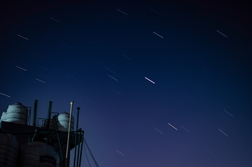 A long exposure photograph featuring streaks of stars moving across a night sky. In the lower portion, there are silo-like structures and cylindrical tanks with piping visible against the dark sky. The vast dark blue sky occupies most of the image.