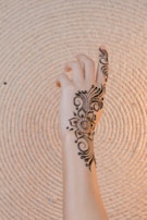 A hand with intricate henna design, featuring floral and swirl patterns. The design extends from the wrist to the fingers, with delicate details emphasizing the beauty of the artwork. The background consists of a textured, spiral-patterned surface.