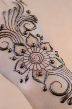 An intricate henna design featuring floral patterns and swirling lines is applied on a forearm. The central motif is a flower with petals radiating around a circular center, complemented by decorative dots and flowing curves.