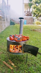A metal stove is situated outdoors on a grassy area, with a small stack of burning firewood inside. Several red bell peppers are being roasted on top of the stove, with some smoke escaping from a chimney pipe. In the background, there are stone walls, a partially visible house, and some trees.
