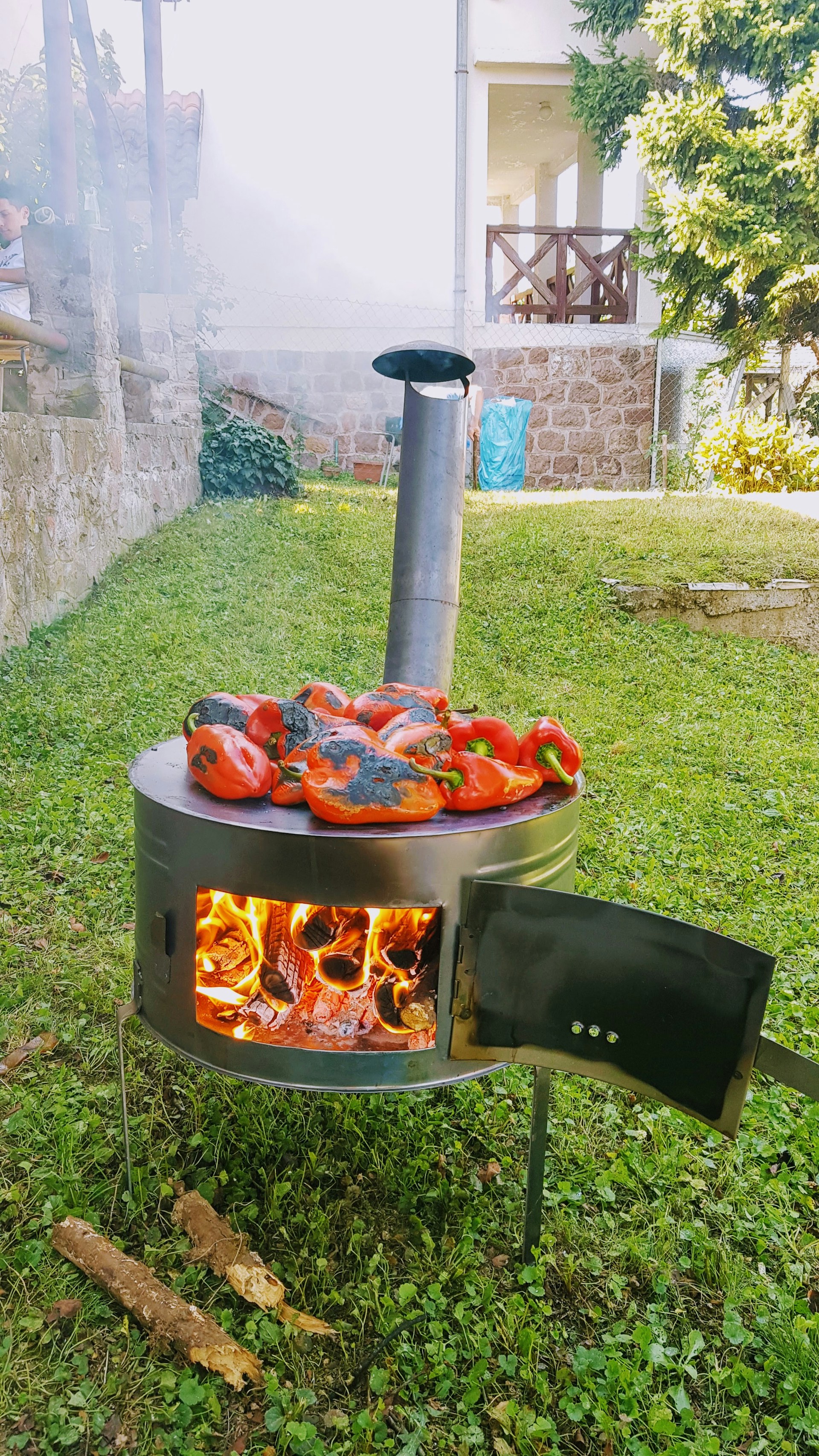 A metal stove is situated outdoors on a grassy area, with a small stack of burning firewood inside. Several red bell peppers are being roasted on top of the stove, with some smoke escaping from a chimney pipe. In the background, there are stone walls, a partially visible house, and some trees.