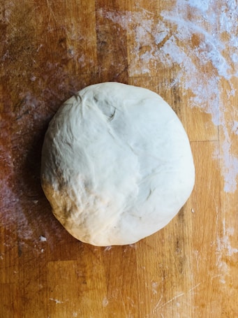 A ball of dough rests on a wooden surface dusted with flour. The light and shadow create a slightly textured look on the dough.