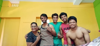 Five people are gathered closely together, smiling at the camera. They stand in front of a brightly colored wall, with yellow and green hues. Two individuals are shirtless, while others wear casual attire. The group seems to be enjoying a moment of camaraderie.
