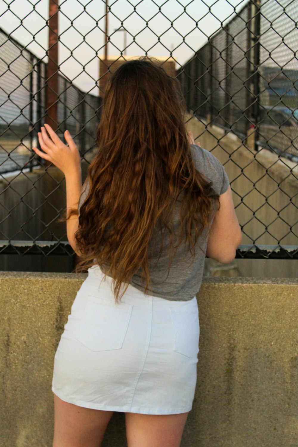 woman wearing white denim skirt and gray shirt holding on black cyclone fence
