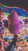 A large pink cotton candy held up in front of a colorful carnival stall adorned with plush toys and bright, multicolored lights.