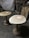 Two wooden tables with natural wood grain patterns on their surfaces are positioned on a rough concrete floor. A large standing fan is in the background, along with various woodworking tools and materials. The setting resembles a workshop or studio with a partially visible canvas or tarp hanging on the wall.