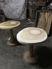 Two wooden tables with natural wood grain patterns on their surfaces are positioned on a rough concrete floor. A large standing fan is in the background, along with various woodworking tools and materials. The setting resembles a workshop or studio with a partially visible canvas or tarp hanging on the wall.