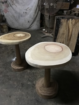 Two wooden tables with natural wood grain patterns on their surfaces are positioned on a rough concrete floor. A large standing fan is in the background, along with various woodworking tools and materials. The setting resembles a workshop or studio with a partially visible canvas or tarp hanging on the wall.