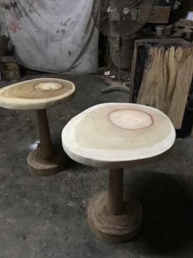 Two wooden tables with natural wood grain patterns on their surfaces are positioned on a rough concrete floor. A large standing fan is in the background, along with various woodworking tools and materials. The setting resembles a workshop or studio with a partially visible canvas or tarp hanging on the wall.