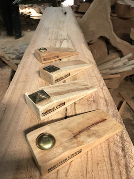 A woodworking setting with four handcrafted wooden doorstops featuring metal accents. Each piece is engraved with the text 'metnis Living'. The background shows a workspace with unfinished wood and shavings.
