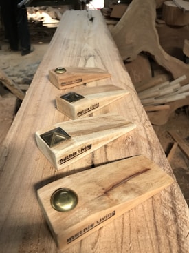 A woodworking setting with four handcrafted wooden doorstops featuring metal accents. Each piece is engraved with the text 