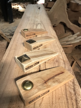 A woodworking setting with four handcrafted wooden doorstops featuring metal accents. Each piece is engraved with the text 'metnis Living'. The background shows a workspace with unfinished wood and shavings.