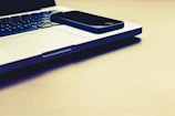 A black smartphone is placed on the closed silver laptop, which is on a light-colored surface. The keyboard of the laptop is visible, and the lighting gives the scene a warm tone.