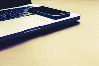 A black smartphone is placed on the closed silver laptop, which is on a light-colored surface. The keyboard of the laptop is visible, and the lighting gives the scene a warm tone.