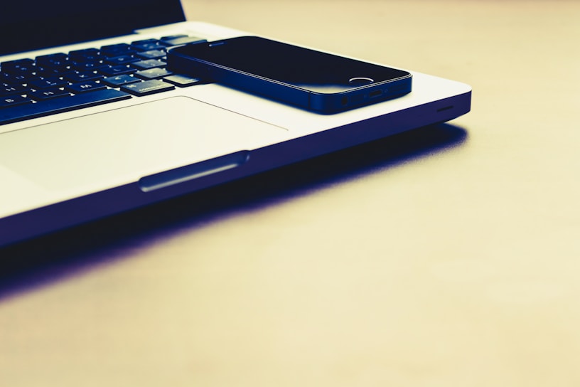 A black smartphone is placed on the closed silver laptop, which is on a light-colored surface. The keyboard of the laptop is visible, and the lighting gives the scene a warm tone.