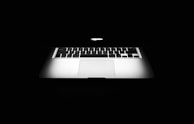 A partially opened laptop with a glowing apple logo on the back of the screen, set against a black background. The keyboard and touchpad are visible, with light reflecting off the metallic surface, creating a sleek and modern appearance.
