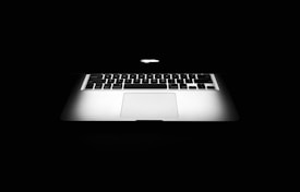 A partially opened laptop with a glowing apple logo on the back of the screen, set against a black background. The keyboard and touchpad are visible, with light reflecting off the metallic surface, creating a sleek and modern appearance.