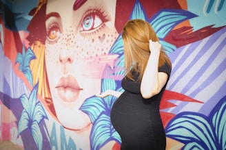 A pregnant woman in a black dress stands in front of a vibrant mural featuring a large, colorful face with striking eyes and freckles. The mural includes abstract leaf patterns and bold geometric shapes in shades of blue, orange, red, and purple.