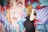 A pregnant woman in a black dress stands in front of a vibrant mural featuring a large, colorful face with striking eyes and freckles. The mural includes abstract leaf patterns and bold geometric shapes in shades of blue, orange, red, and purple.