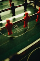 A foosball table is shown with red and black player figures positioned on the rods. The surface features the UEFA Champions League logo, indicating a themed table design.