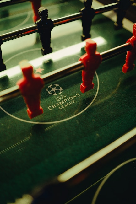A foosball table is shown with red and black player figures positioned on the rods. The surface features the UEFA Champions League logo, indicating a themed table design.