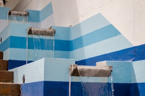 A series of cascading water features set against a wall with blue and white geometric patterns. The water flows smoothly from metal spouts set in light blue concrete structures. The scene is tidy and structured, with a modern and minimalist aesthetic.