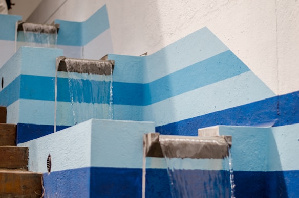 A series of cascading water features set against a wall with blue and white geometric patterns. The water flows smoothly from metal spouts set in light blue concrete structures. The scene is tidy and structured, with a modern and minimalist aesthetic.