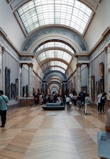 An indoor hall of a museum with high, vaulted ceilings topped with a series of skylights. The hall's walls are adorned with various artworks, and it is supported by decorative marble columns. Visitors, some standing, walking, and others sitting on a central bench, are scattered throughout the space, engaging with the art and each other.