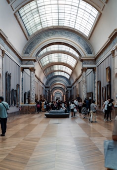 An indoor hall of a museum with high, vaulted ceilings topped with a series of skylights. The hall's walls are adorned with various artworks, and it is supported by decorative marble columns. Visitors, some standing, walking, and others sitting on a central bench, are scattered throughout the space, engaging with the art and each other.