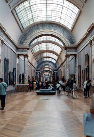 An indoor hall of a museum with high, vaulted ceilings topped with a series of skylights. The hall's walls are adorned with various artworks, and it is supported by decorative marble columns. Visitors, some standing, walking, and others sitting on a central bench, are scattered throughout the space, engaging with the art and each other.