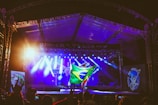 A music concert stage is brightly lit with blue and white spotlights, featuring a person holding a large Brazilian flag at the center. The event appears festive, with visible audience members in the foreground and large screens flanking the stage.