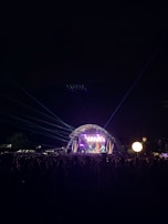 A large outdoor concert stage is illuminated with bright lights and lasers, set against the dark night sky. A large crowd of people stands in front of the stage, enjoying the performance. The stage features a patterned arch structure, and colorful lighting enhances the visual atmosphere.