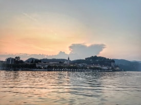 A serene lakeside scene at sunset with a quaint town in the foreground and hills in the background. The sky is tinged with warm hues as the sun sets, casting reflections on the calm water.