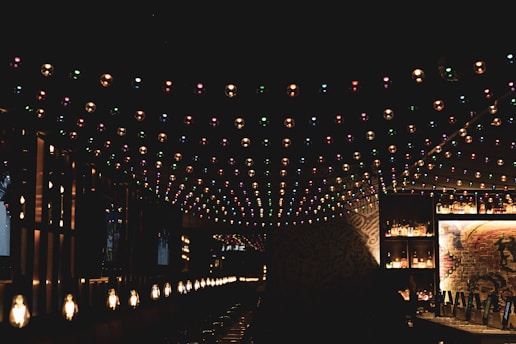 A dimly lit bar or lounge area with colorful string lights hanging from the ceiling. The bar is lined with bottles and has a warm, inviting atmosphere. Soft, yellowish lights are positioned along the counter, casting a cozy glow. The background features some exposed brick with artistic graffiti or murals.