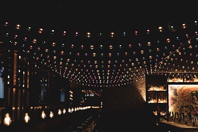 A dimly lit bar or lounge area with colorful string lights hanging from the ceiling. The bar is lined with bottles and has a warm, inviting atmosphere. Soft, yellowish lights are positioned along the counter, casting a cozy glow. The background features some exposed brick with artistic graffiti or murals.