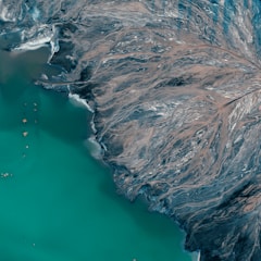 An aerial view of a coastline where a teal body of water meets a rugged landscape streaked with patterns resembling river deltas or veins. This earthy area consists of dark and brown tones, creating intricate, almost abstract patterns against the water.