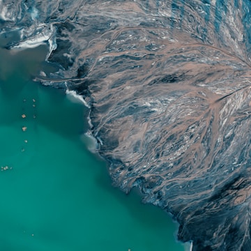 An aerial view of a coastline where a teal body of water meets a rugged landscape streaked with patterns resembling river deltas or veins. This earthy area consists of dark and brown tones, creating intricate, almost abstract patterns against the water.