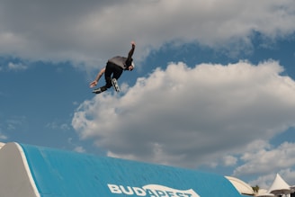 A person wearing inline skates performs an acrobatic jump against a backdrop of a partly cloudy blue sky. The lower part of the image shows a blue ramp with the word 'BUDAPEST' on it.